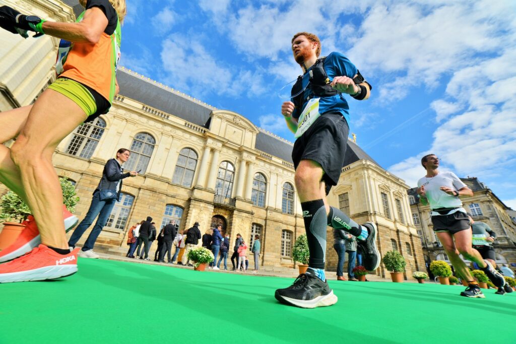 Des coureurs participant à une course à pied à Rennes passant le palais du Parlement de Bretagne sous un ciel bleu. En arrière-plan des spectateurs observent l’événement sportif. Les athlètes portent des tenues et équipements de course, et le sol est recouvert d'un tapis vert vif pour baliser le parcours.