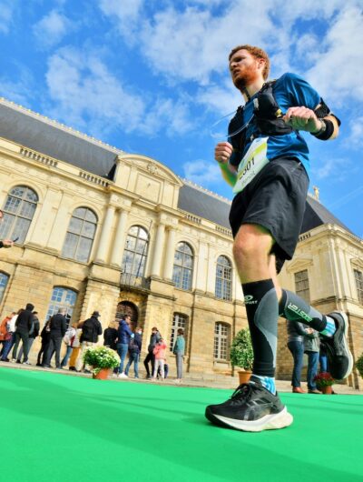 Des coureurs participant à une course à pied à Rennes passant le palais du Parlement de Bretagne sous un ciel bleu. En arrière-plan des spectateurs observent l’événement sportif. Les athlètes portent des tenues et équipements de course, et le sol est recouvert d'un tapis vert vif pour baliser le parcours.