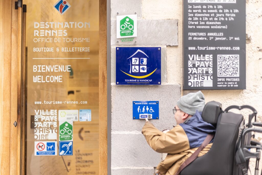 Une personne en fauteuil roulant devant l'entrée de l'Office de tourisme de Rennes. Destination Rennes est labellisée Tourisme & Handicap